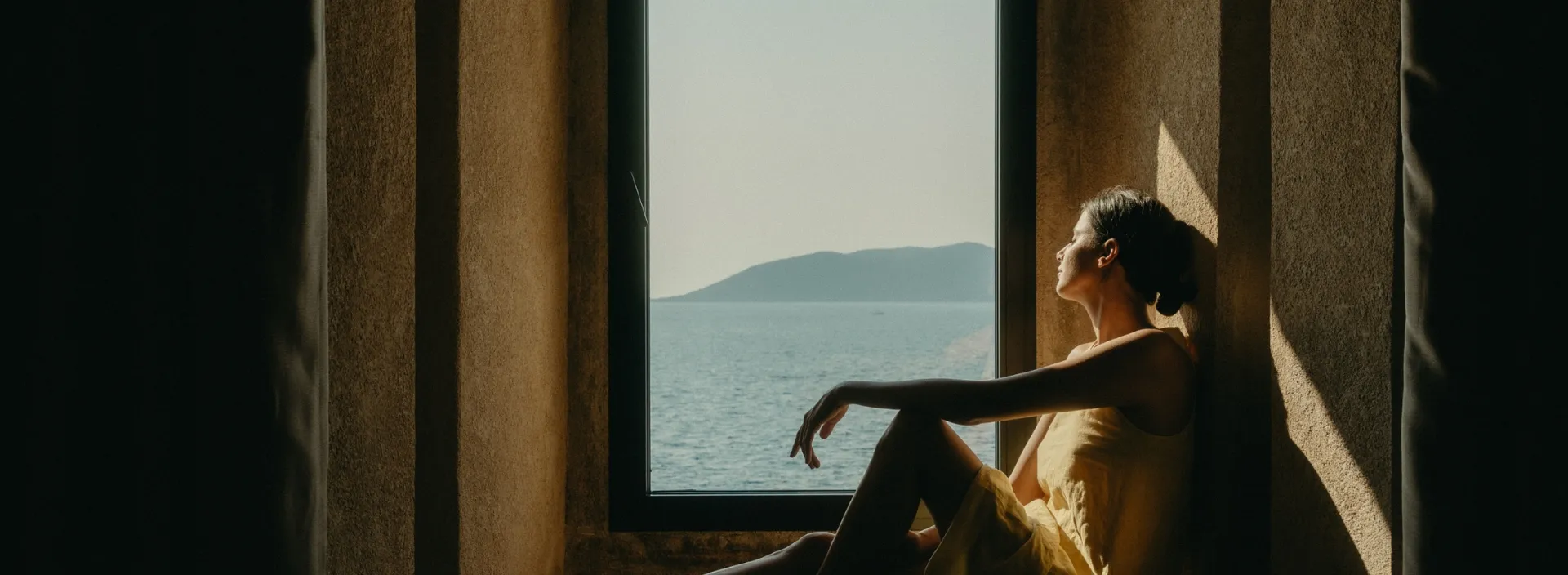 A woman rests against a window sill, bathed in warm light inside Mamula Island by Banyan Tree.